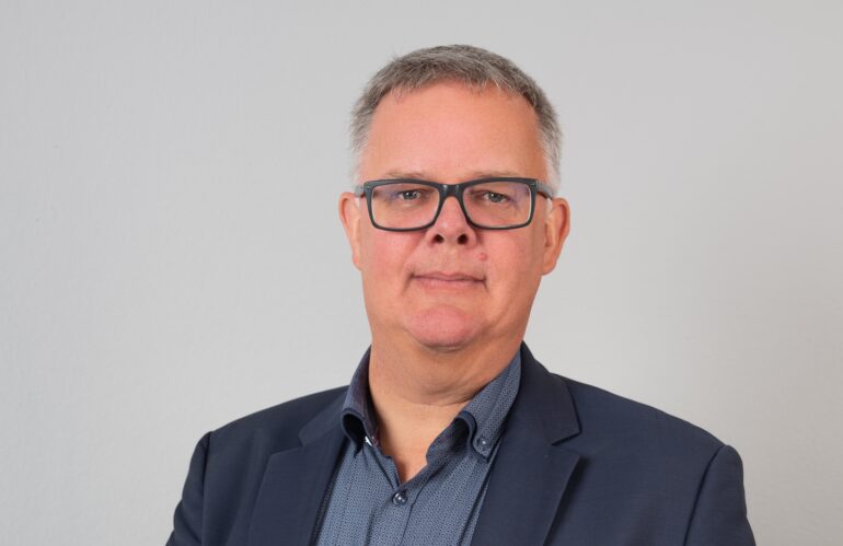 Ein Mann mit Brille posiert für ein Foto in einer Unternehmensumgebung.